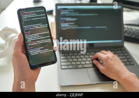 Close-up of programmer holding smartphone with codes in her hands and connecting it with laptop at table Stock Photo