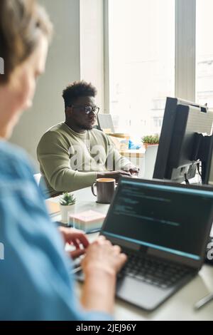 African young man sitting at table in front of computer monitor and typing with his colleague sitting opposite him and working on laptop Stock Photo