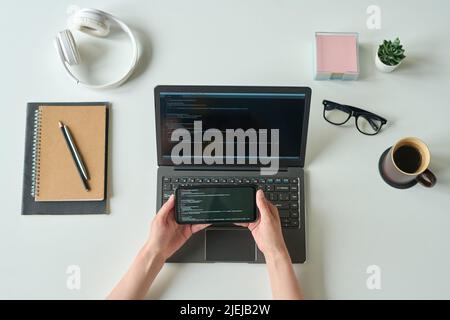 Above view of programmer connecting smartphone with codes with laptop at her workplace Stock Photo