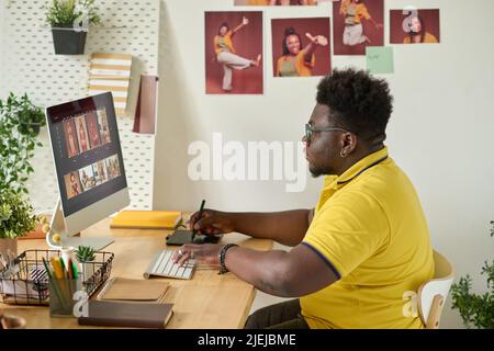 African retoucher sitting at his workplace and using graphic tablet to retouch photo on computer screen at office Stock Photo