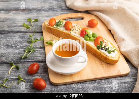 Long white bread sandwich with cream cheese, tomatoes and microgreen on gray wooden background and linen textile. side view, close up. Stock Photo
