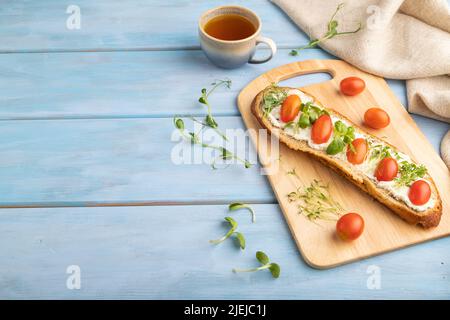 Long white bread sandwich with cream cheese, tomatoes and microgreen on blue wooden background and linen textile. side view, copy space. Stock Photo