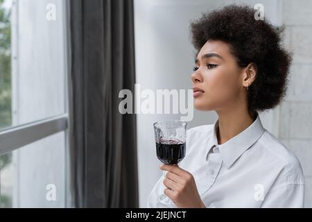 upset african american woman holding glass of red wine and looking at window Stock Photo