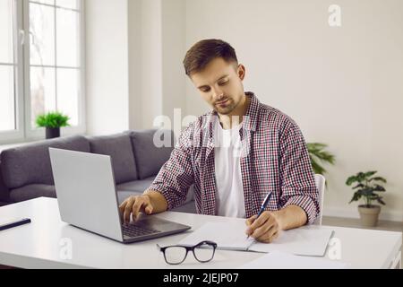 Serious student sitting at desk, using his laptop computer and writing in notebook Stock Photo