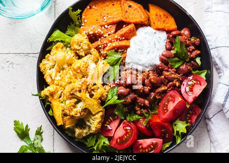 Baked vegetables buddha bowl, close-up. Cooked sweet potato, cauliflower, tomato and bean salad, top view. Vegan recipe. Stock Photo