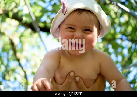 Mother holds in the air a cute happy baby who smiles in front of the branches of a tree with green leaves. Summer sunny day. Happiness concept. Stock Photo