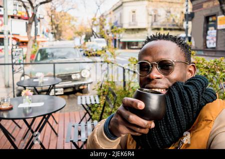 young african man takes selfie while drinking coffee at, sitting outdoors outside restaurant, city in background with copy space. Stock Photo