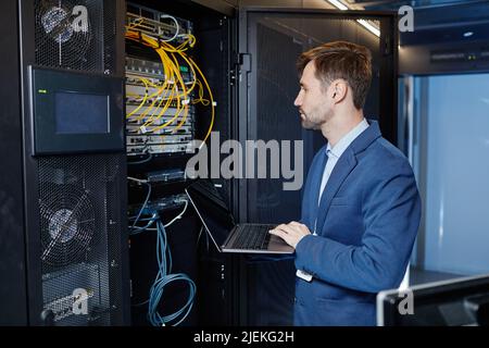 Side view portrait of Caucasian young man holding laptop while setting up internet network in server room Stock Photo