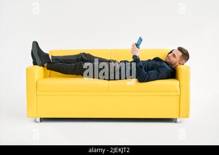 Relaxed man lying on yellow sofa using smartphone over white background Stock Photo