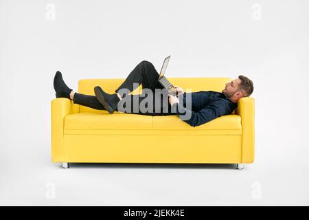 Relaxed man lying on yellow sofa using laptop over white background Stock Photo