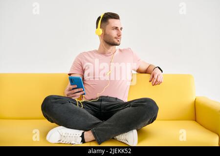 Relaxed young man wear headphones listen music by his mobile phone while sitting on yellow sofa over white background Stock Photo