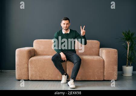 Trendy man showing hand gesture V sign for victory or peace looking ro camera while sitting on modern apartment Stock Photo