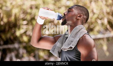 Athlete drinking water from a bottle during an outdoor workout. Fit, athletic african american man taking break to hydrate and refresh while standing Stock Photo