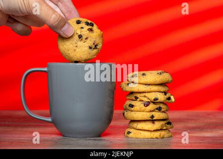stack of home cooked cookies and a grey mug against a red background with a hand dunking a cookie. Stock Photo