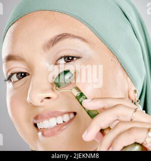 Muslim woman smiling and using a facial roller on her skin. Beauty model doing her morning skin care routine with a jade roller. Young female using a Stock Photo