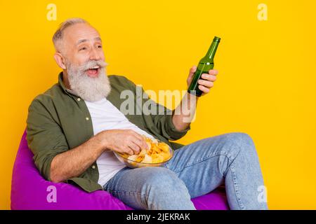 Photo of cheerful bearded man hold beer bottle chips bowl watch football wear green shirt isolated yellow color background Stock Photo