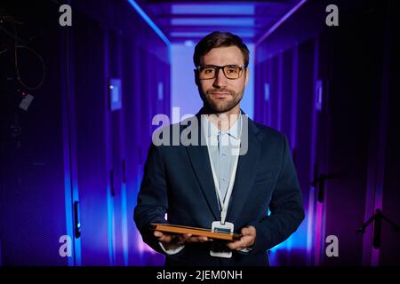 Waist up portrait of male network engineer holding tablet in server room and looking at camera Stock Photo