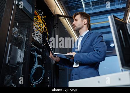 Low angle portrait of IT engineer holding laptop while setting up internet network in server room Stock Photo