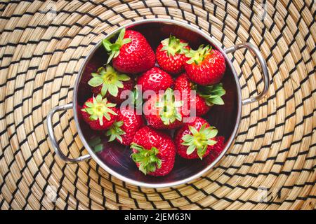 A metallic bowl of red ripe strawberries on a circular straw mat, viewed from above / flat lay Stock Photo