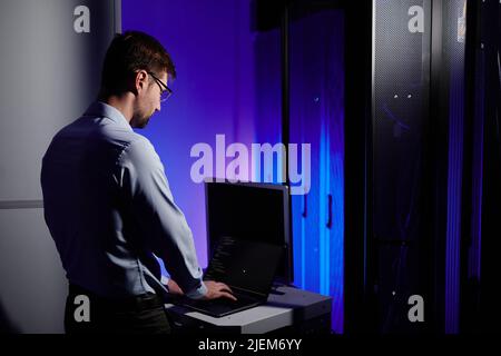 Back view of network engineer using laptop in dark server room lit by neon light, copy space Stock Photo