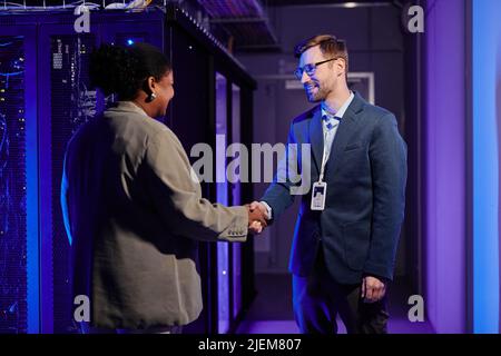 Waist up portrait of two business partners shaking hands while standing in data center interior Stock Photo