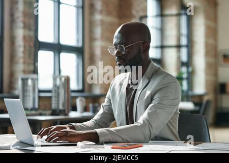 African serious businessman in eyeglasses sitting at his workplace at office and working on laptop Stock Photo
