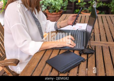 White woman teleworking in the garden with mobile phone and laptop. Stock Photo