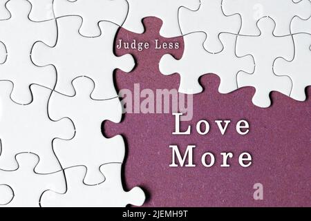 Motivational quote on white jigsaw puzzle with some missing pieces on purple background - Judge less love more. Stock Photo