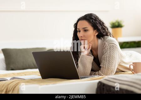 Focused young woman with laptop pc working or studying online, lying on bed at home, copy space Stock Photo