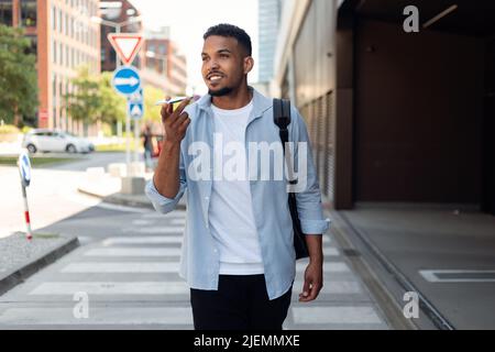 Handsome african american guy recording audio message on mobile phone, having conversation while walking in the city Stock Photo