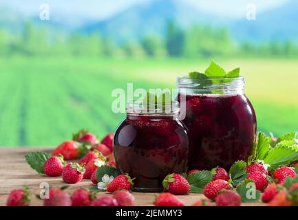 Strawberry jam jars on rustic table. Strawberry garden image in the background. Natural spring jams. Organic breakfast food concept. Stock Photo