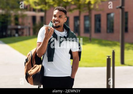 Positive stylish black guy talking on smartphone, using loudspeaker or recording audio message, walking outdoors Stock Photo