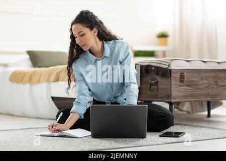 Serious young woman sitting on floor, working on laptop, taking notes in notebook in bedroom Stock Photo