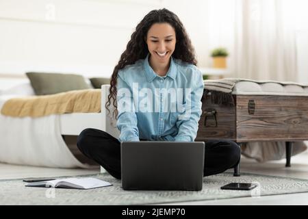 Beautiful young woman sitting on floor, working on laptop pc in bedroom, full length Stock Photo