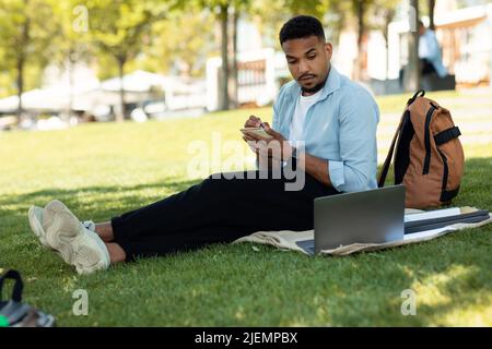 Freelance employee working remote, education concept. Busy black guy with laptop making notes in notepad sitting in park Stock Photo