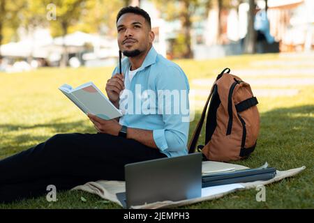 Pensive black male student learning outdoors, writing notes while sitting with laptop nearby on blanket in park Stock Photo
