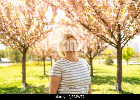 Beautiful cute blonde 50s woman near the tree blossoms with pink flowers, profile, horizontal