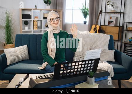 Portrait of muslim girl in hijab playing piano. Pretty women in music therapy by playing piano on music room. Young arab female girl in piano lesson. Relaxing at home waving hand. Music concept. Stock Photo