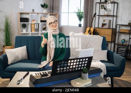 Portrait of muslim girl in hijab playing piano. Pretty women in music therapy by playing piano on music room. Young arab female girl in piano lesson. Relaxing at home waving hand. Music concept. Stock Photo