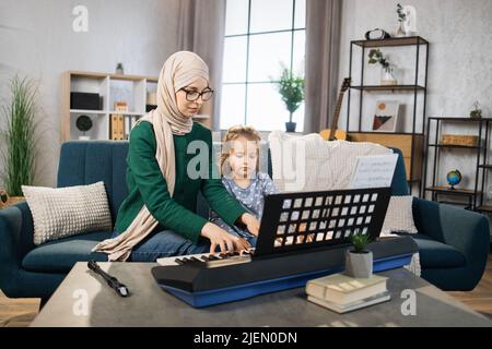 Beautiful young muslim woman in hijab and her charming little daughter are smiling while playing classic digital piano at home. Mother teaching cute little musician girl to play piano. Stock Photo