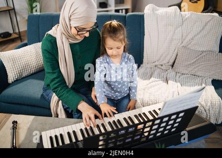 Little girl child learning to play classic digital piano with her young islam mom in hijab at home. Small kid preschool student playing musical instrument with muslim mother. Stock Photo