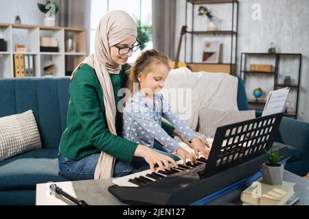 Portrait of muslim teacher in hijab and her small student playing piano at home. Mom and daughter playing musician instrument on background of light living room at home. Stock Photo