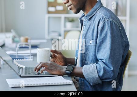 Cropped side view shot of young black man using laptop while working in office and holding cup of coffee, copy space Stock Photo
