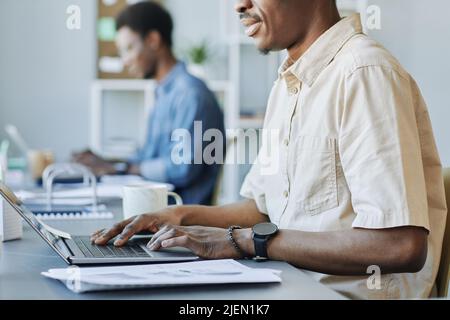 Cropped side view shot of adult black man typing at laptop while working in office and holding cup of coffee, copy space Stock Photo