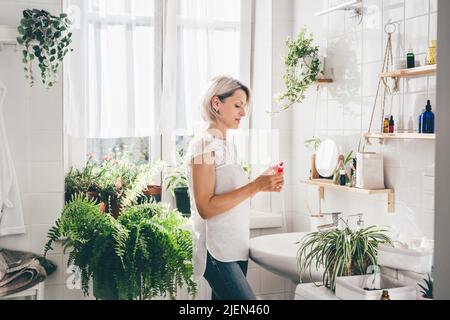 Woman of middle-aged staying in eco friendly white bathroom. Wooden shelves with cosmetics and toiletries against white tile wall. Minimalistic design. Wellness. Comfort home zone Stock Photo