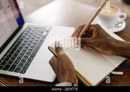 cropped multiracial businessman using laptop, taking notes in workbook, drink coffee in street cafe. High angle Stock Photo