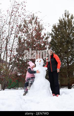 Low angle of smiling middle aged woman with daughter hugging snowman on ground in daytime while having walk in park with trees in background. Parents Stock Photo
