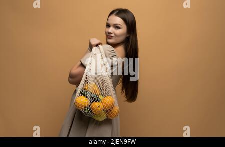 Woman in beige dress holding string shopping reusable mesh bag with vegetables, fruits on a shoulder. Zero waste concept. Space for text. Reusable eco Stock Photo