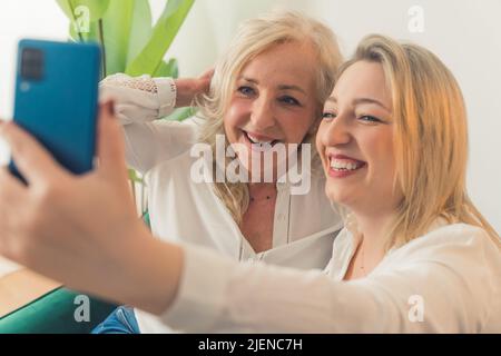 Close-up indoor shot of two good-looking pretty blonde caucasian cheerful women taking a selfie sitting on a sofa and laughing. High quality photo Stock Photo