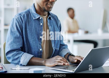Cropped shot of smiling black man working in office and using laptop in minimal grey interior, copy space Stock Photo